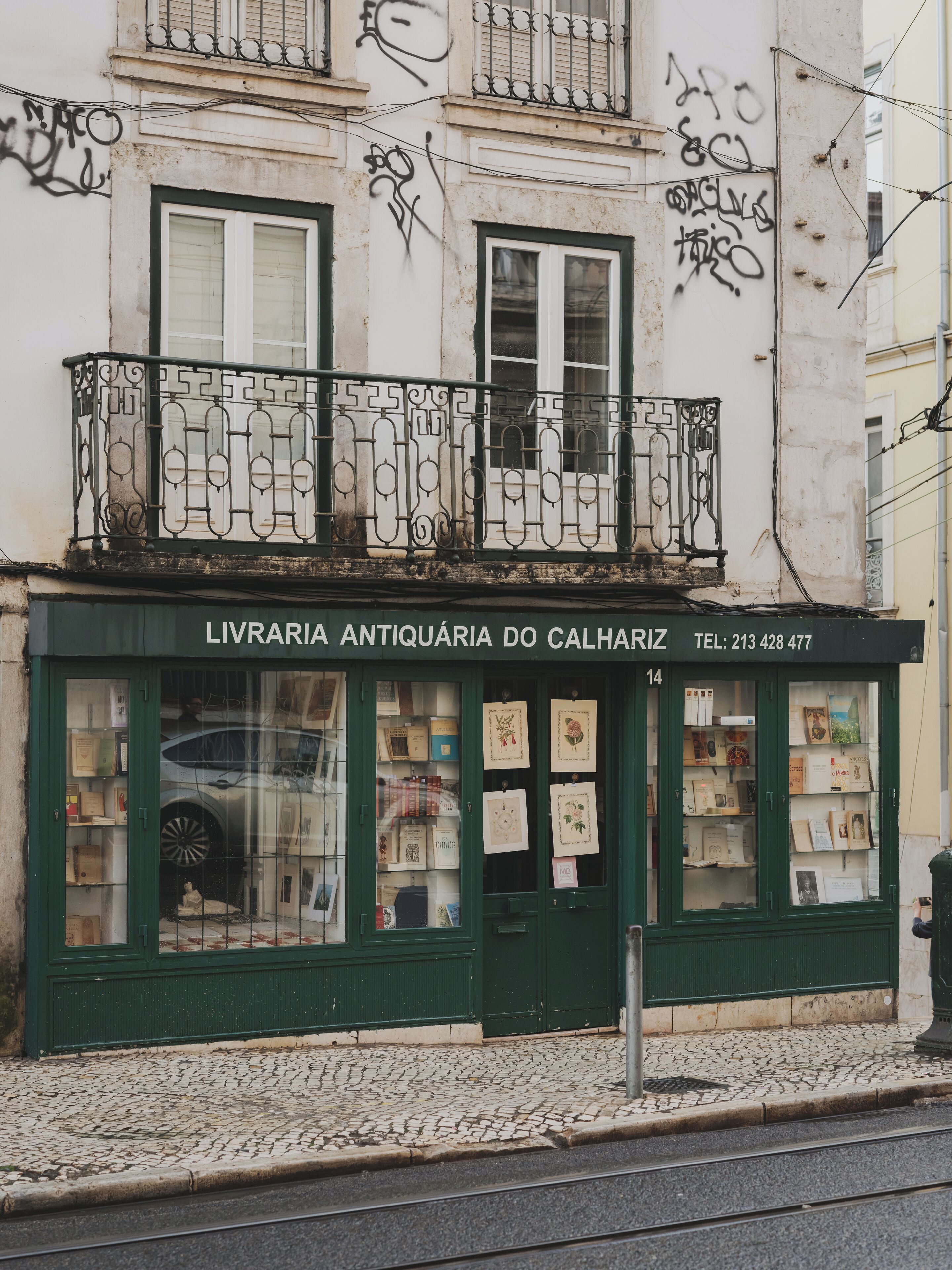 Une librairie à la façade verdoyante, de grandes vitrines où sont exposés des livres, et un balcon agrémenté de balustrades en métal ouvragé.