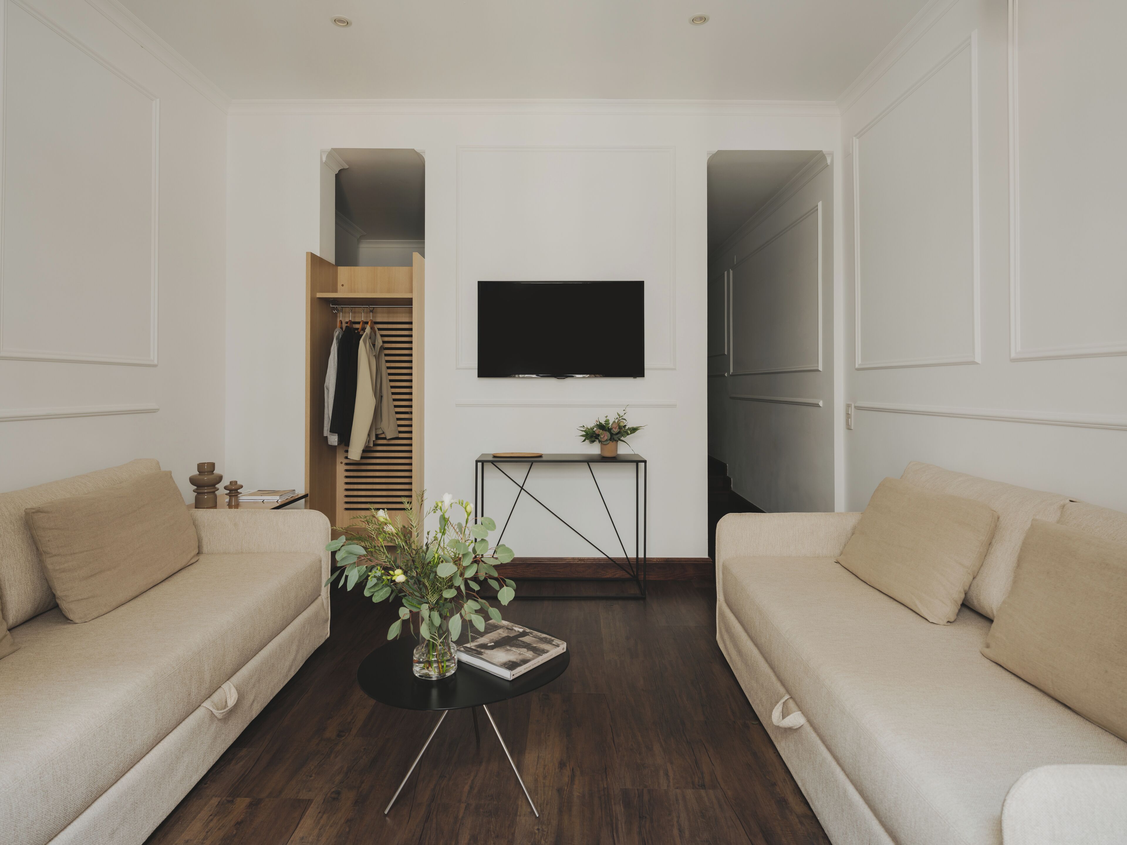 Un salon moderne avec deux canapés beige, une table basse noire ornée d’un vase, un téléviseur à écran plat fixé au mur et une armoire en bois où sont suspendus des vêtements.