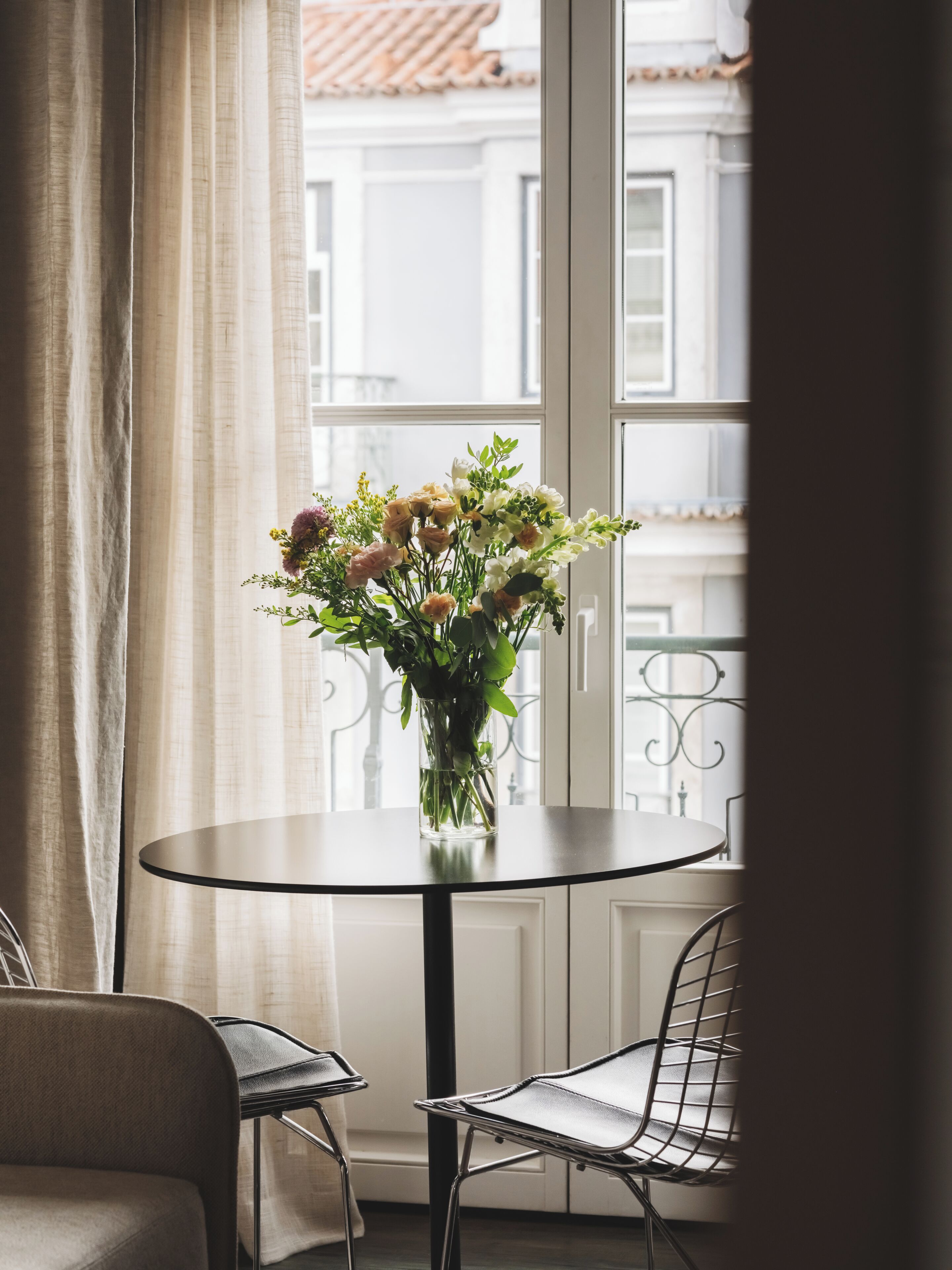 Ett runt bord med en vas med blommor i ett rum med utsikt över en byggnad genom ett fönster.
