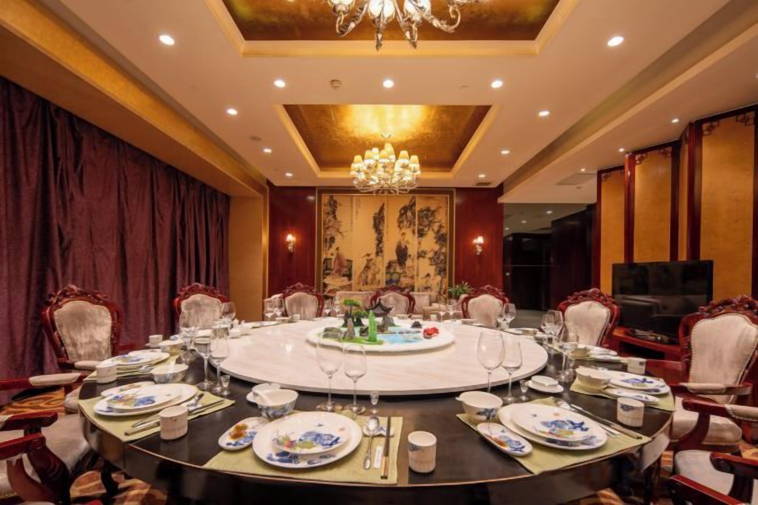 A round dining table set for a meal with chairs around it, a chandelier above, and a television on the wall.