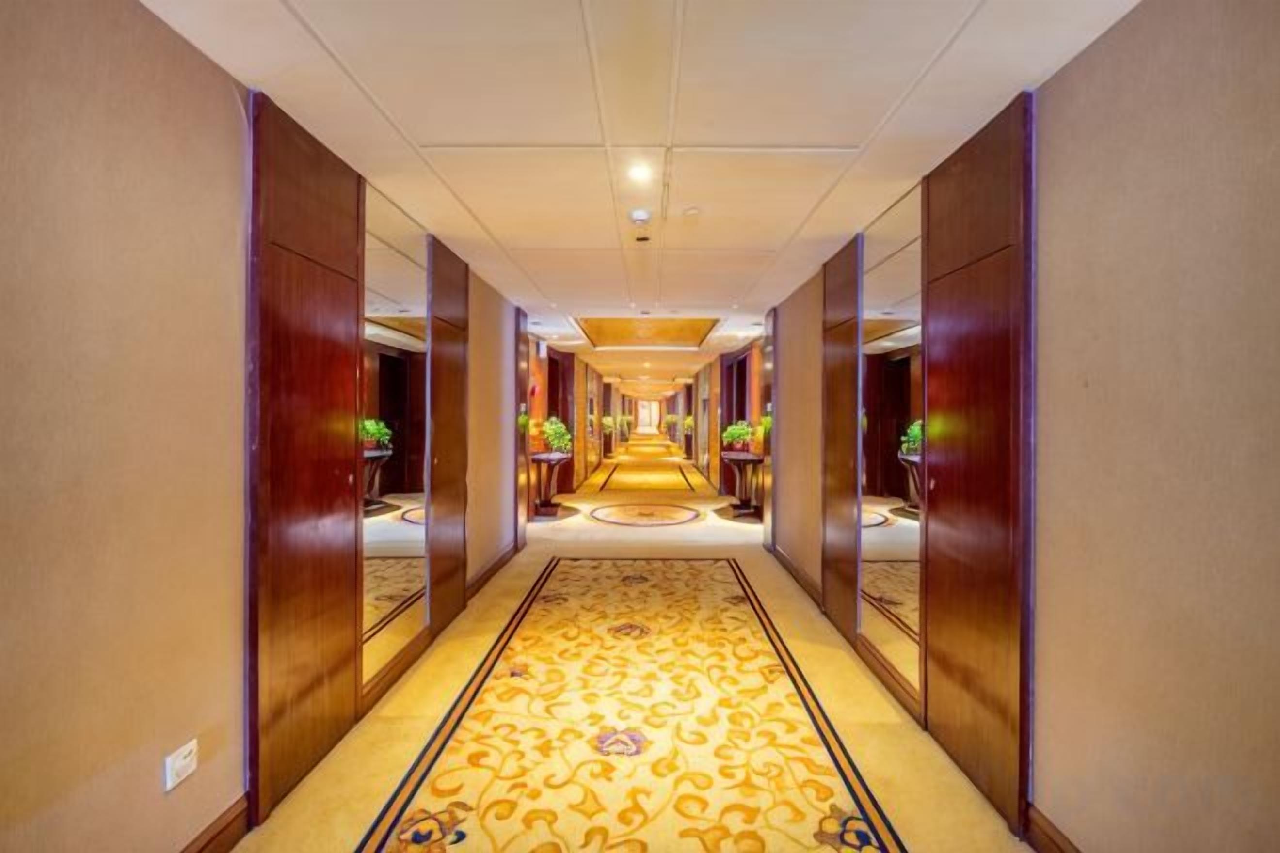 A hotel corridor with a carpeted floor, mirrored walls, and a central light fixture.