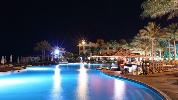 Une piscine nocturne aux bords illuminés, bordée de palmiers, avec un bâtiment à l’architecture traditionnelle en arrière-plan.