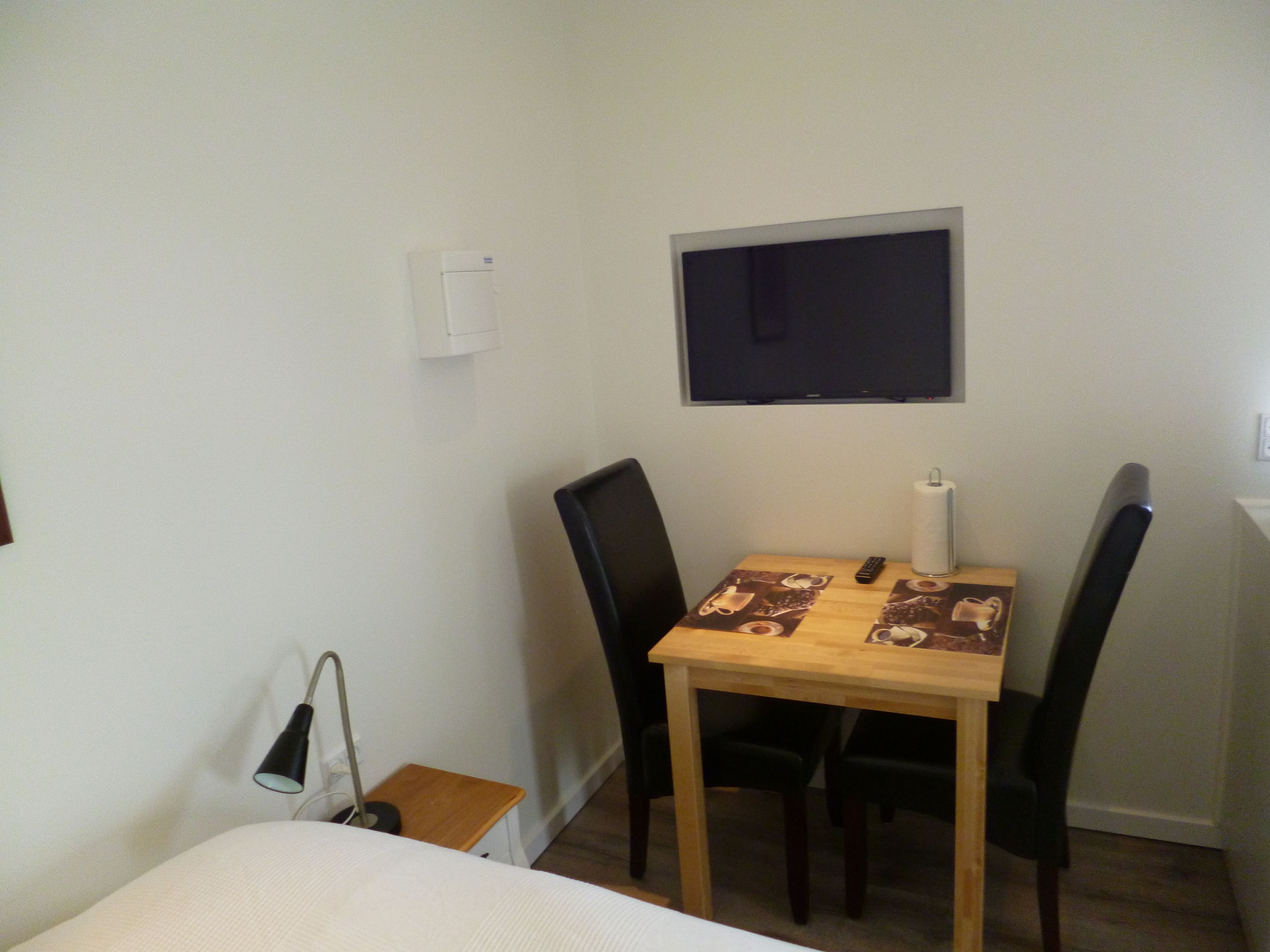 Una habitación pequeña y sencilla con una cama, una mesa con sillas, un televisor montado en la pared y un calentador.
