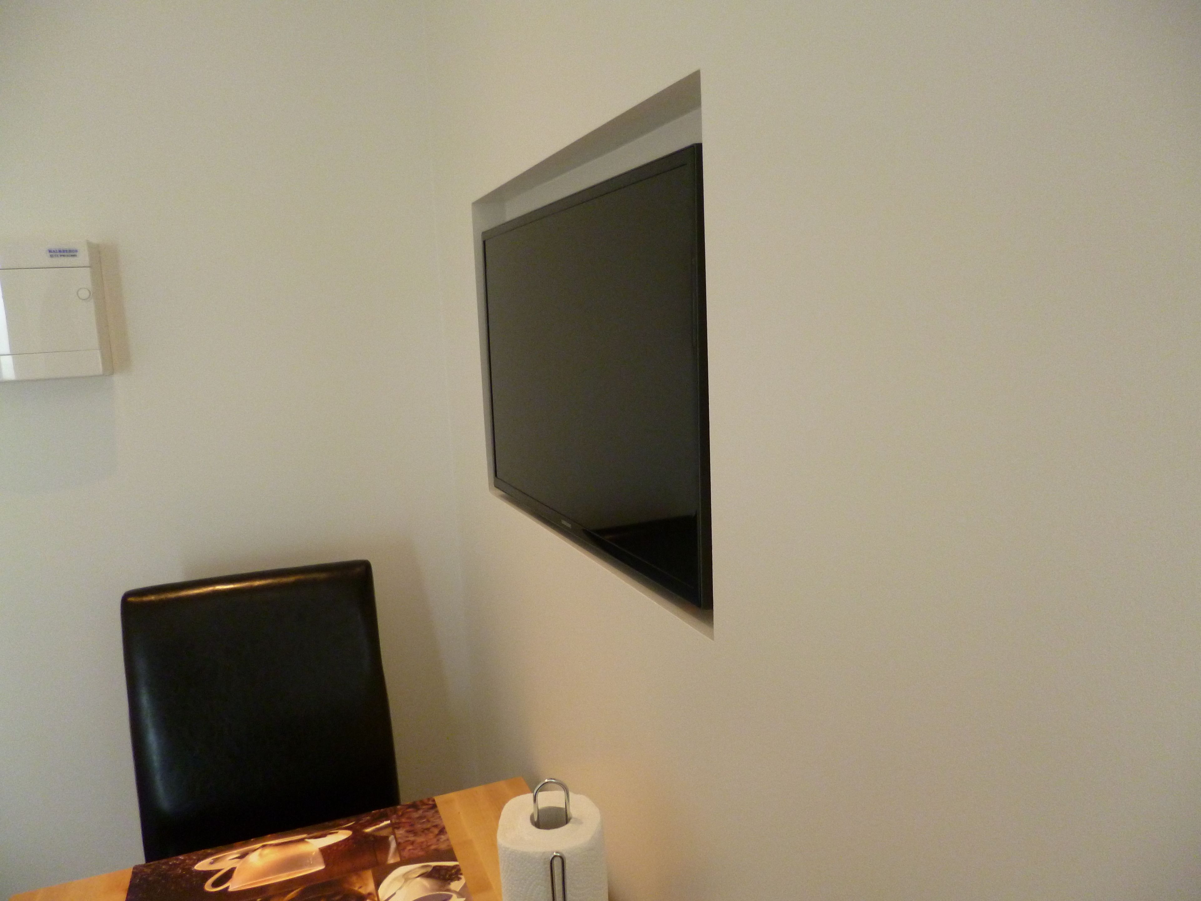 A room with a flat-screen TV mounted on the wall, a black chair, a table with a roll of toilet paper, and a white air conditioning unit.