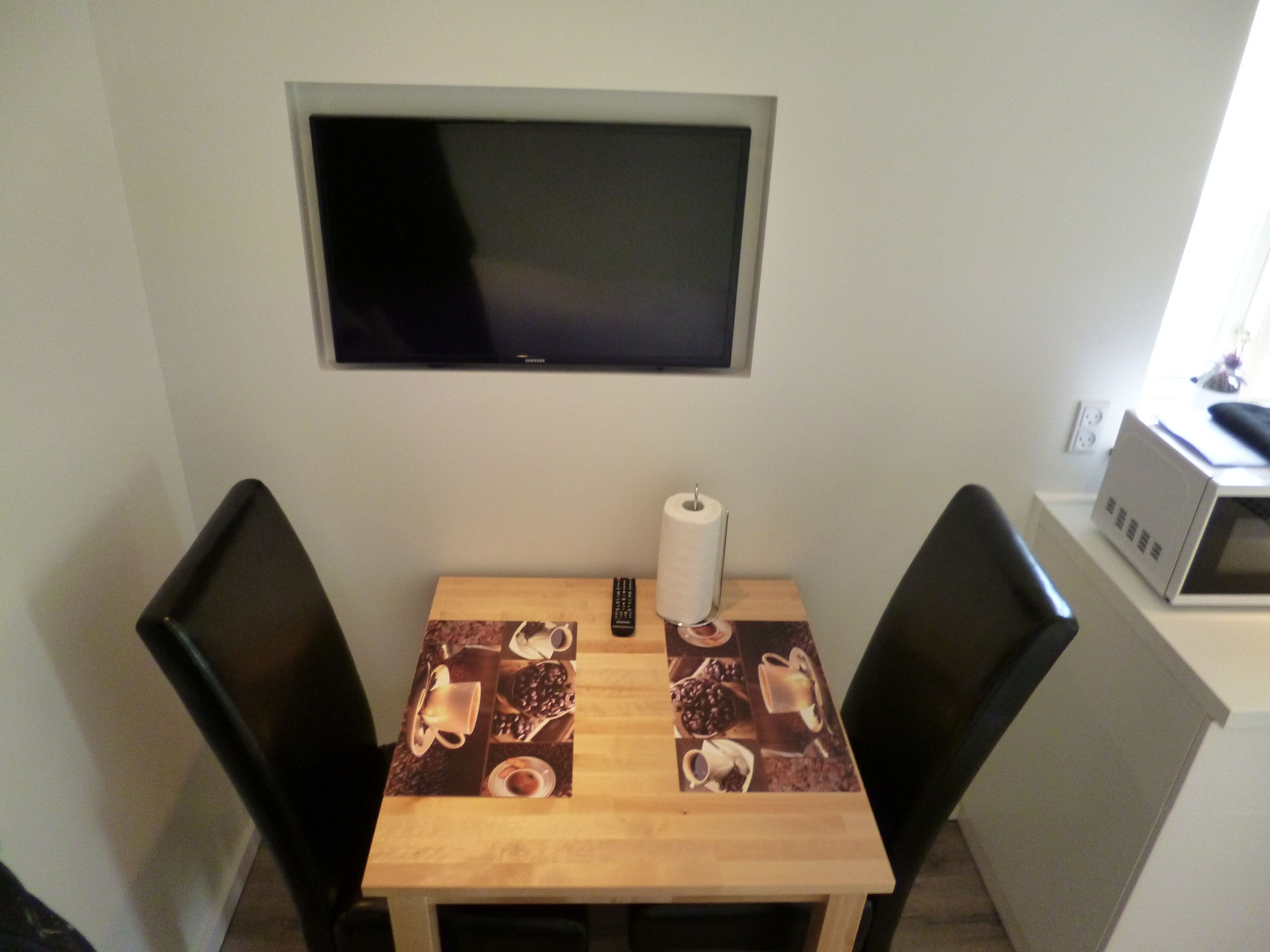 A small dining area with a table, chairs, a remote control, and a TV mounted on the wall.
