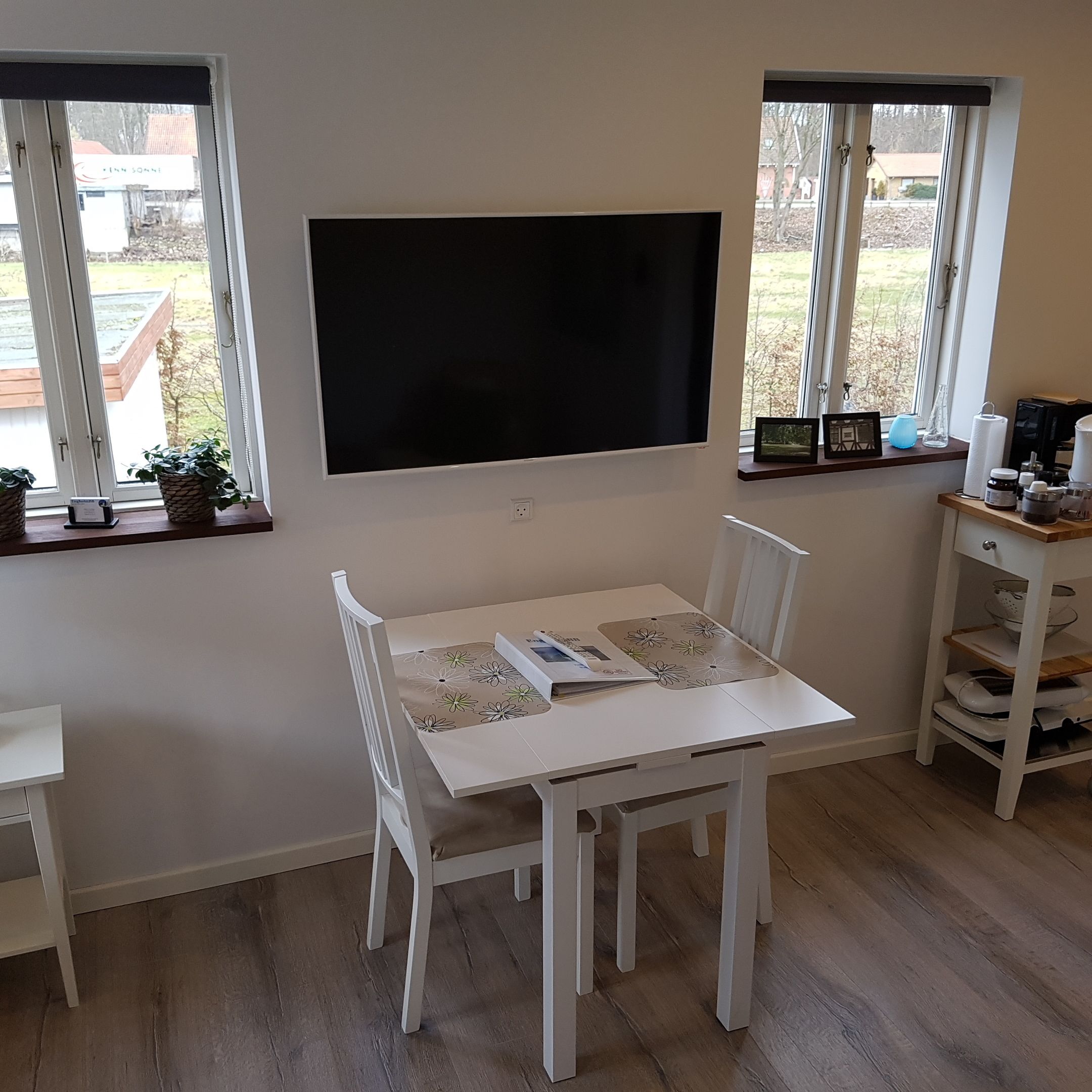 A white dining table with chairs, a flat-screen TV mounted on the wall, and a window with a view of a garden.