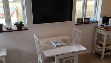 A white dining table with chairs, a flat-screen TV mounted on the wall, and a window with a view of a garden.