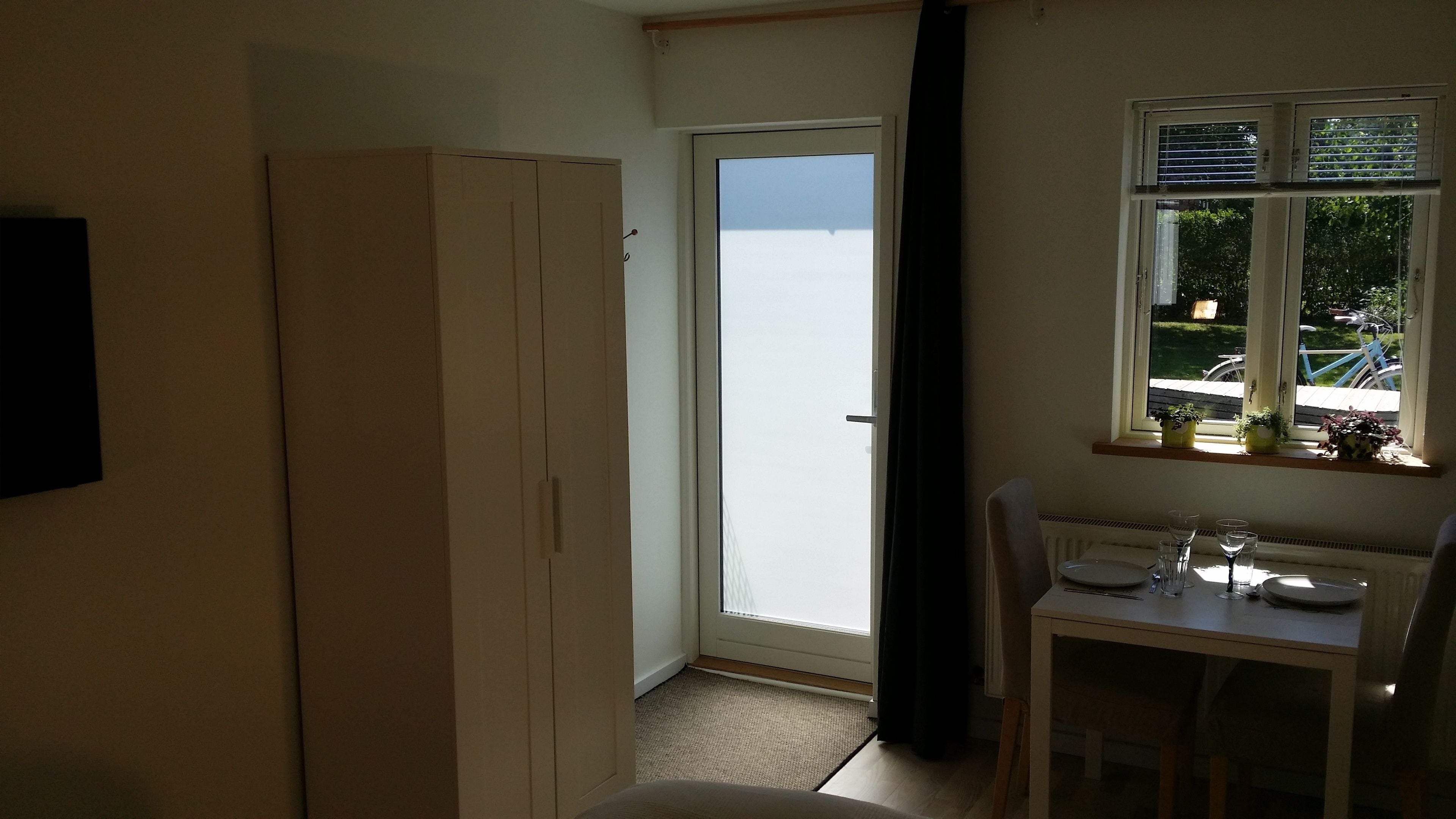 A small room with a white wardrobe, a table with a chair, a window with blinds, and a view of the outside.