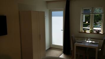 A small room with a white wardrobe, a table with a chair, a window with blinds, and a view of the outside.