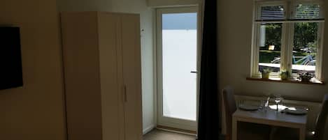 A small room with a white wardrobe, a table with a chair, a window with blinds, and a view of the outside.