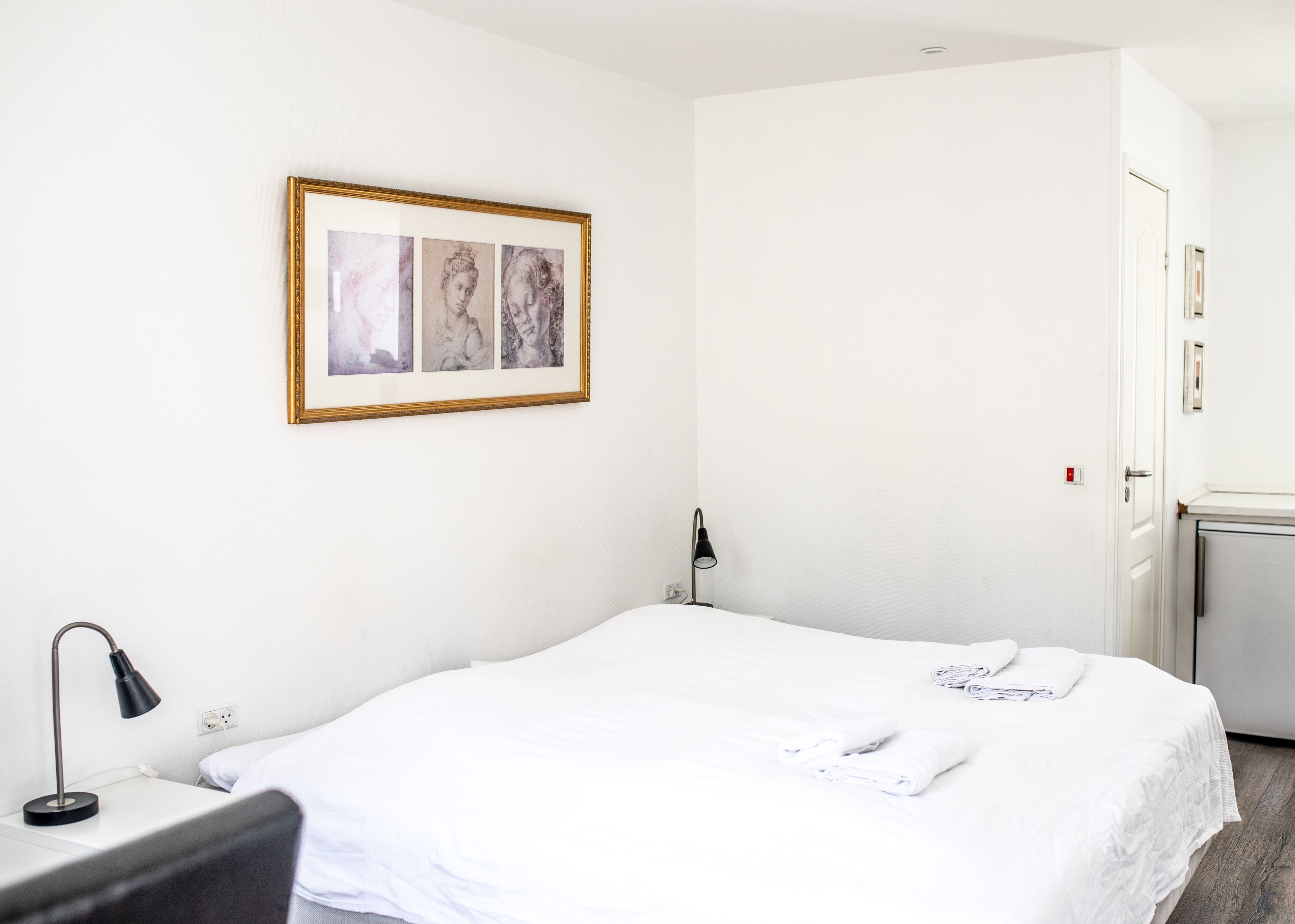 A hotel room with a bed, a desk with a lamp, and a door.