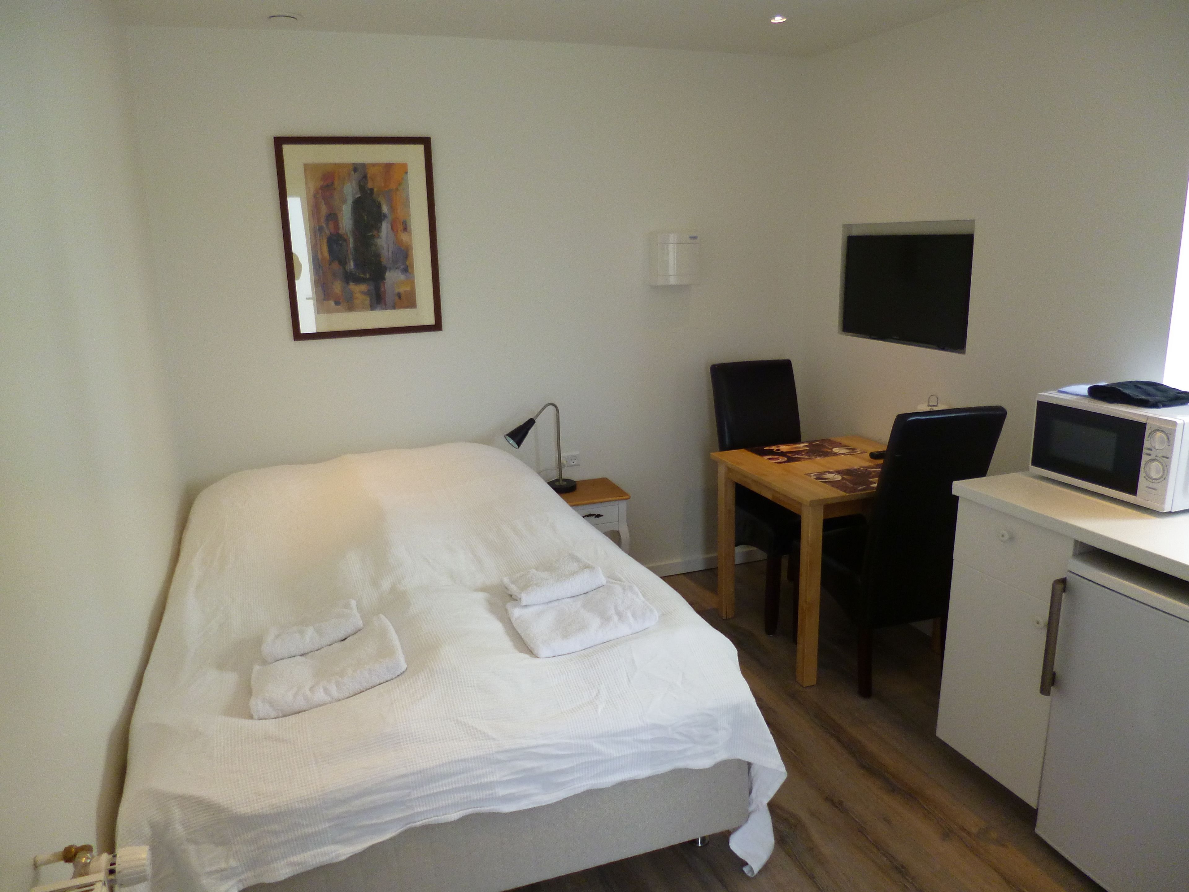 Una habitación pequeña y ordenada con una cama, un escritorio con una computadora, un microondas y una silla.