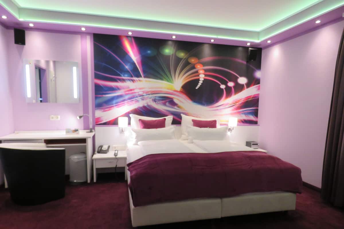 A modern hotel room with a large bed, bedside tables, a vanity, and a colorful abstract mural on the wall.