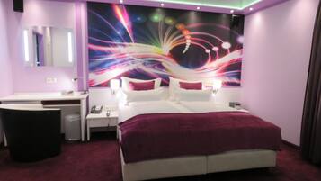 Une chambre d’hôtel moderne, dotée d’un grand lit, de tables de chevet, d’une coiffeuse et d’une fresque murale abstraite et colorée.