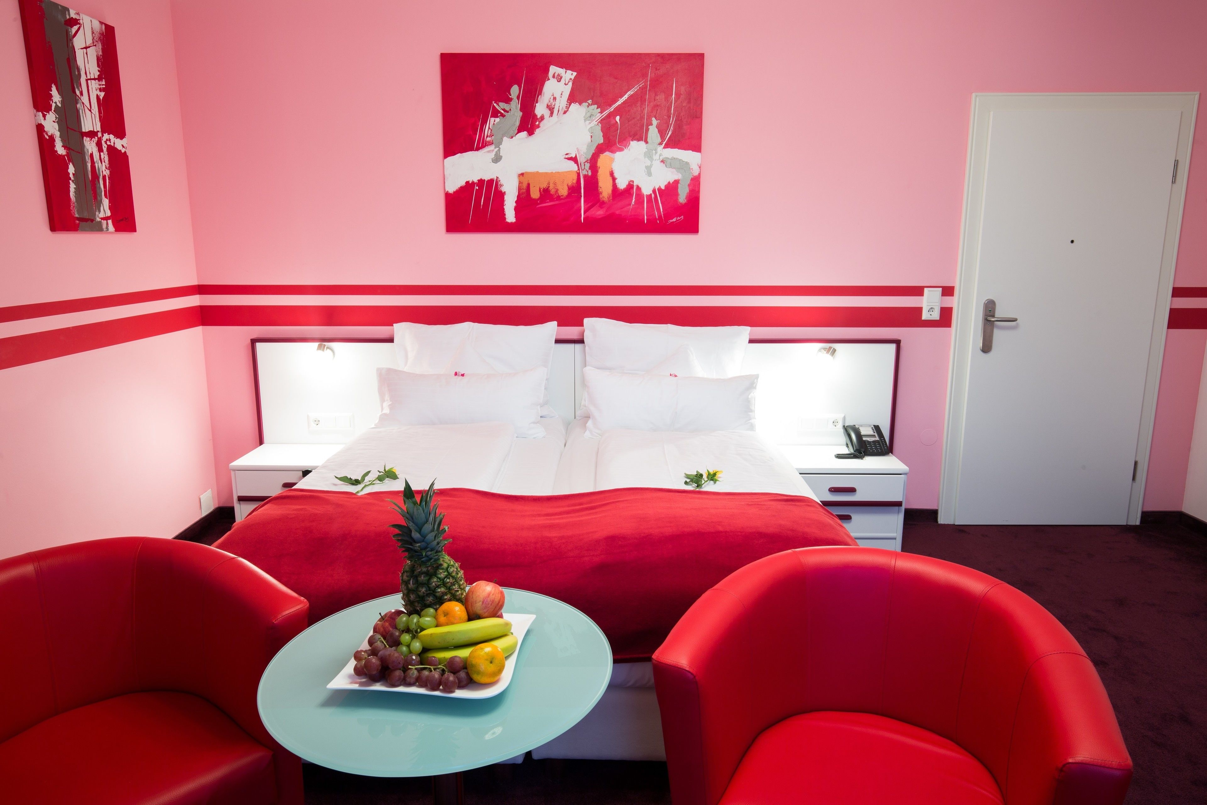 Una habitación de hotel con una cama, dos mesitas de noche, una obra de arte con marco rojo en la pared, un frutero sobre una pequeña mesa y sillas rojas.