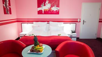 Una habitación de hotel con una cama, dos mesitas de noche, una obra de arte con marco rojo en la pared, un frutero sobre una pequeña mesa y sillas rojas.