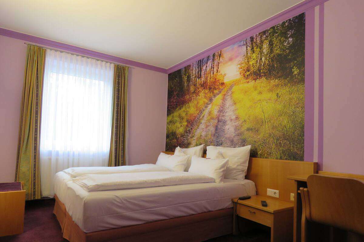 A double bed with white linens, a nightstand, a chair, and a large forest-themed wall mural.