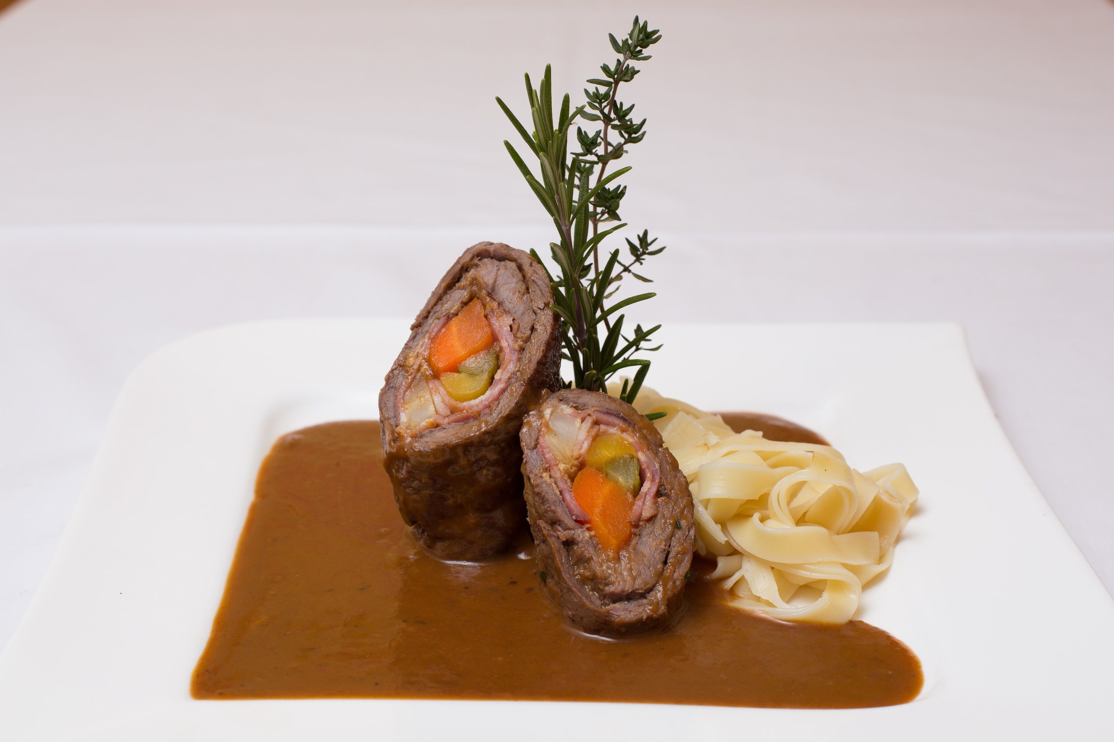 A plate with rolled meat, vegetables, and pasta, garnished with a sprig of rosemary.
