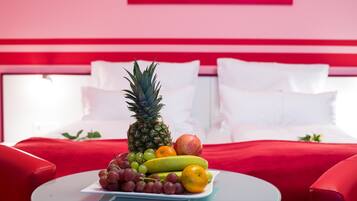 A bed with white pillows and a red headboard, a small round table with a fruit plate in front, and a red couch to the left.