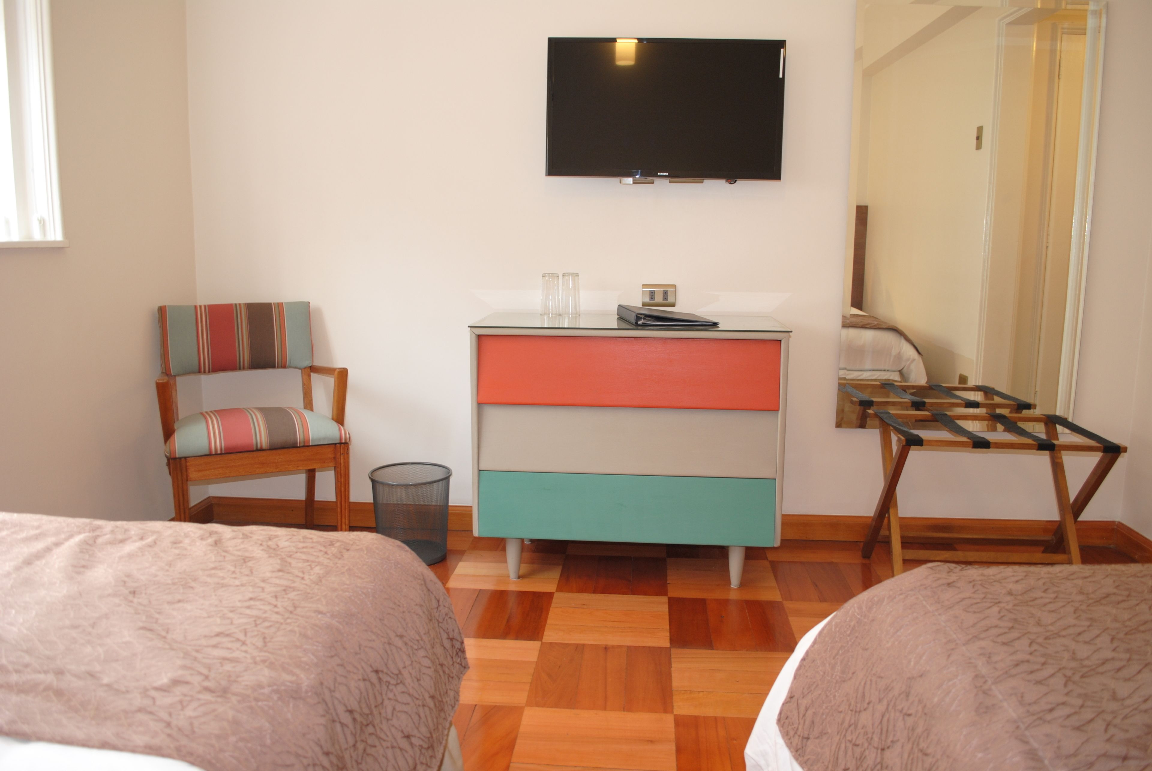 A hotel room with two beds, a television mounted on the wall, a small table with glasses, and a chair with a striped cushion.