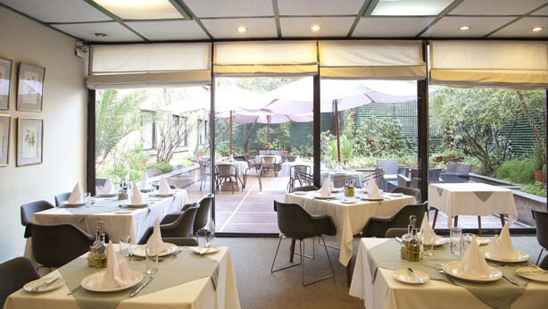 An indoor dining area with tables set for service, large windows, and a view of the outdoor garden. - Hotel Santa Lucia (Santiago)