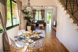 Uma área de refeições com mesa de vidro, cadeiras brancas e um sofá. Há pratos de comida, copos e taças sobre a mesa.