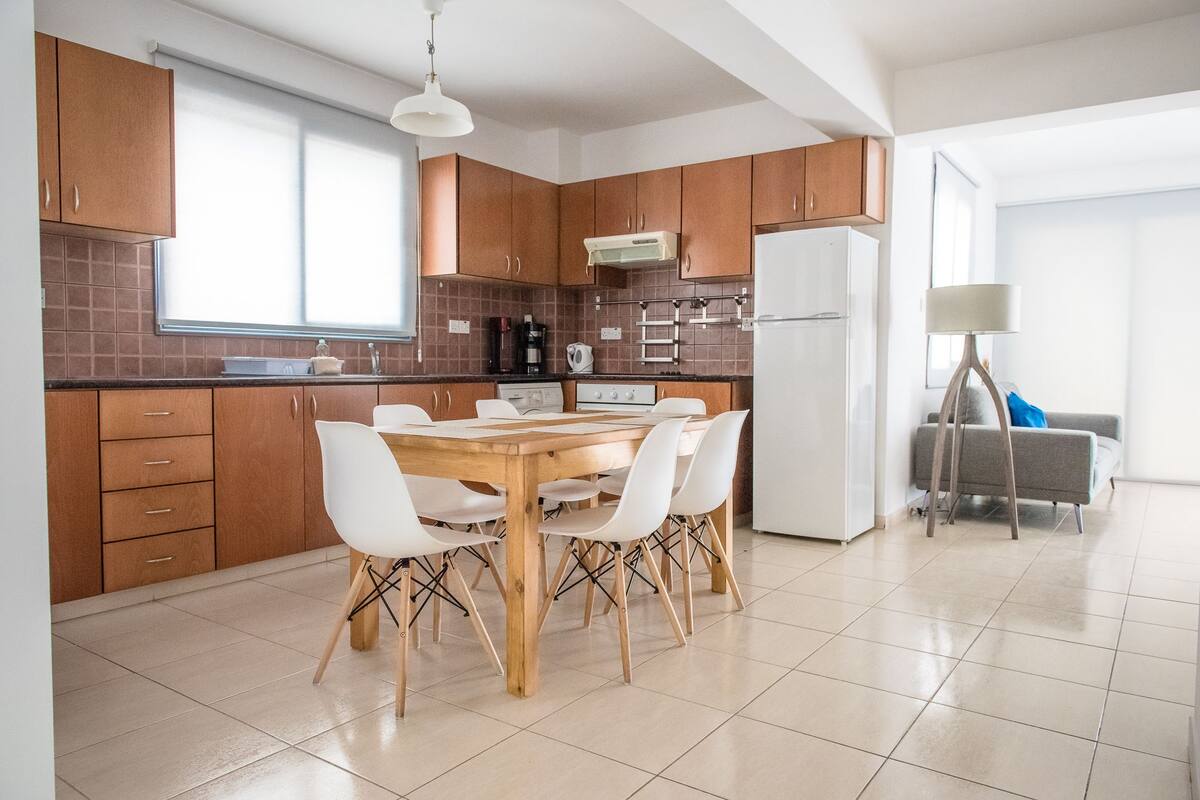 A modern kitchen with wooden cabinets, a dining table with white chairs, a refrigerator, and a lamp.