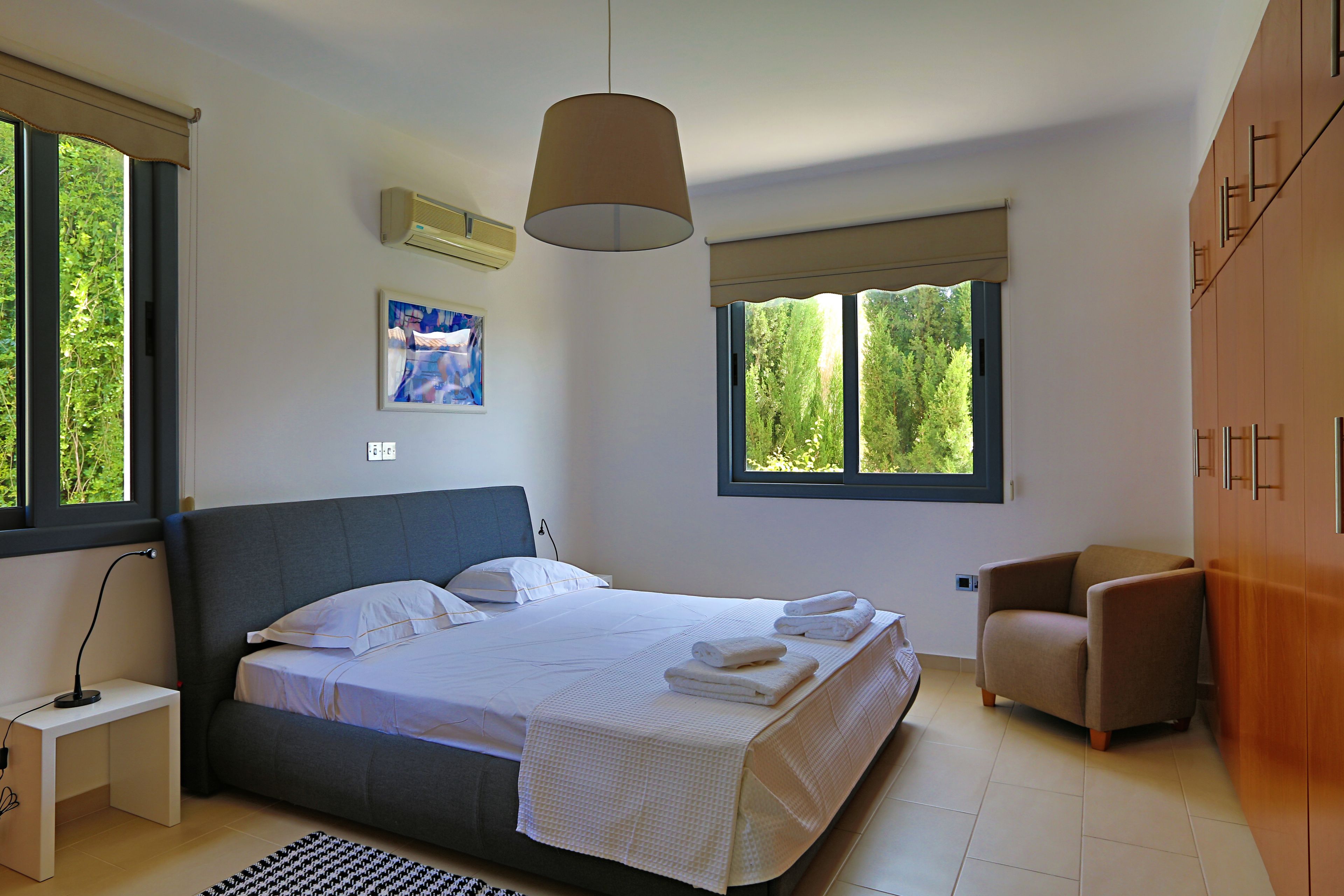 A modern bedroom with a large bed, a bedside table, a chair, and a window with a view of greenery.