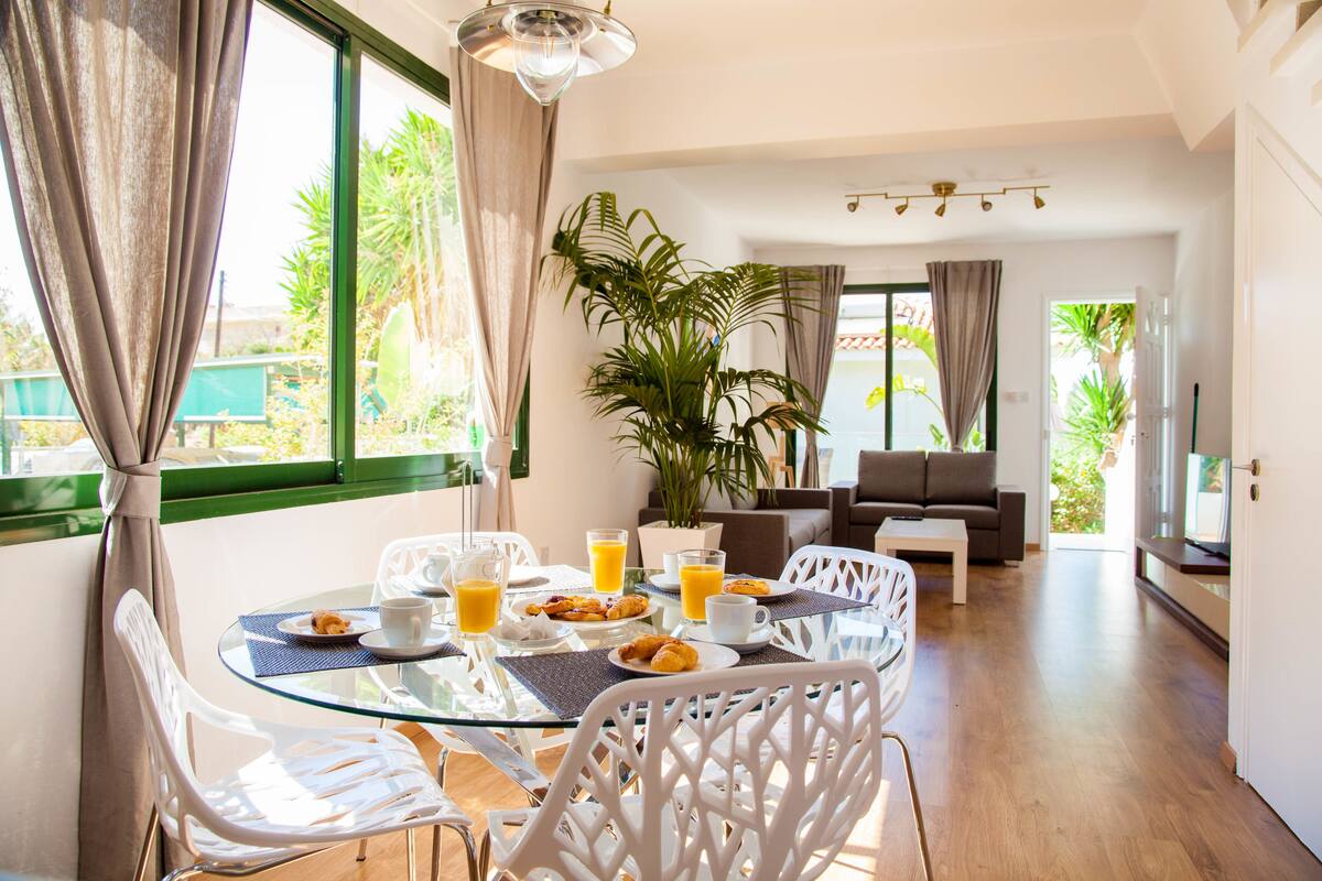 A dining area with a glass table, white chairs, and breakfast items. There is a sofa and a large plant in the background.