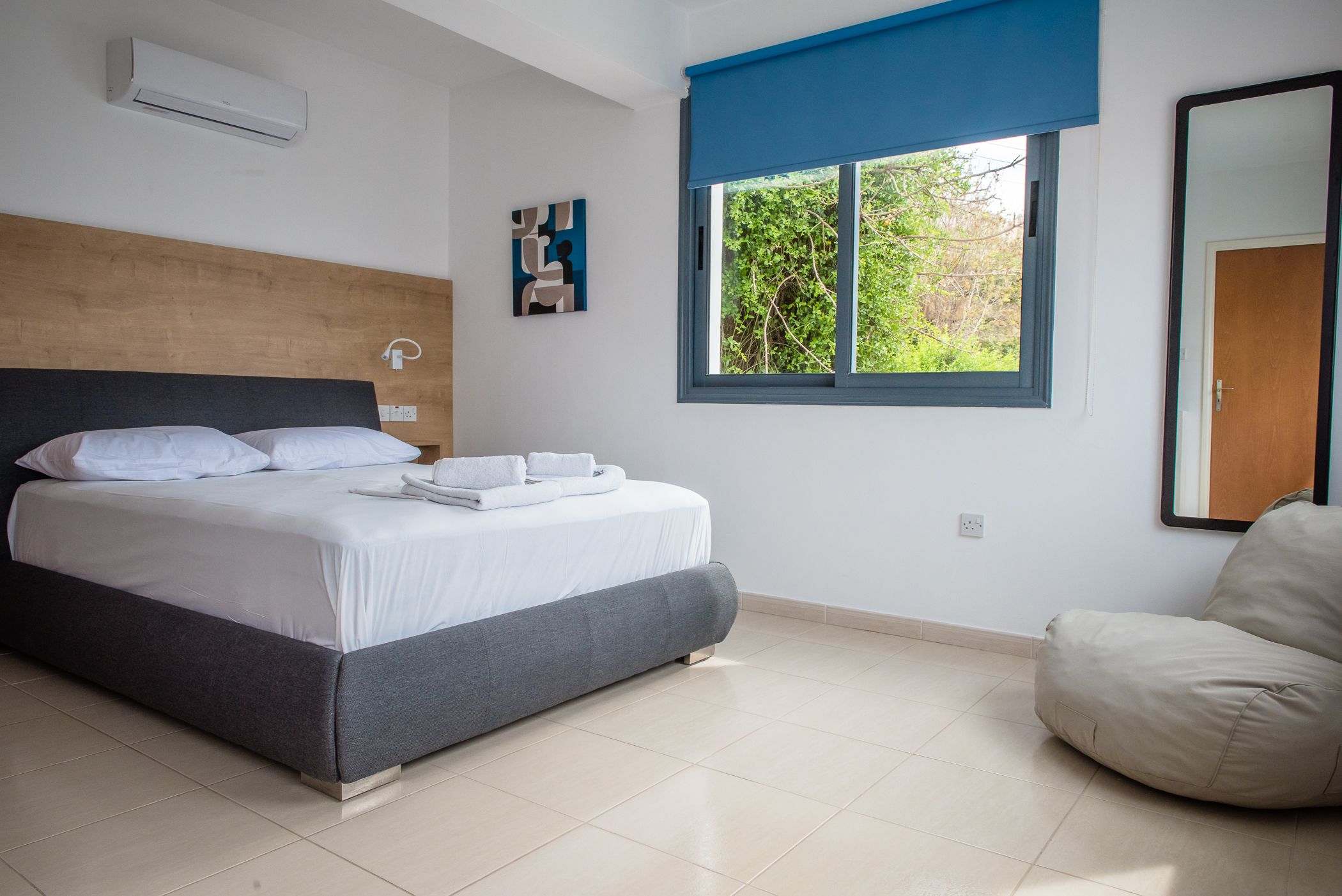 A modern bedroom with a bed, a window with blue blinds, a chair, and a wall-mounted air conditioner.