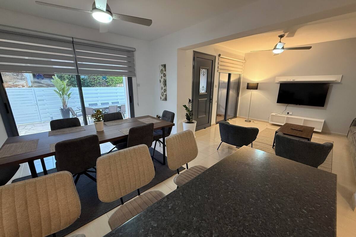 A modern living room with a dark marble coffee table, a flat-screen TV mounted on the wall, and a dining area with a wooden table and chairs.