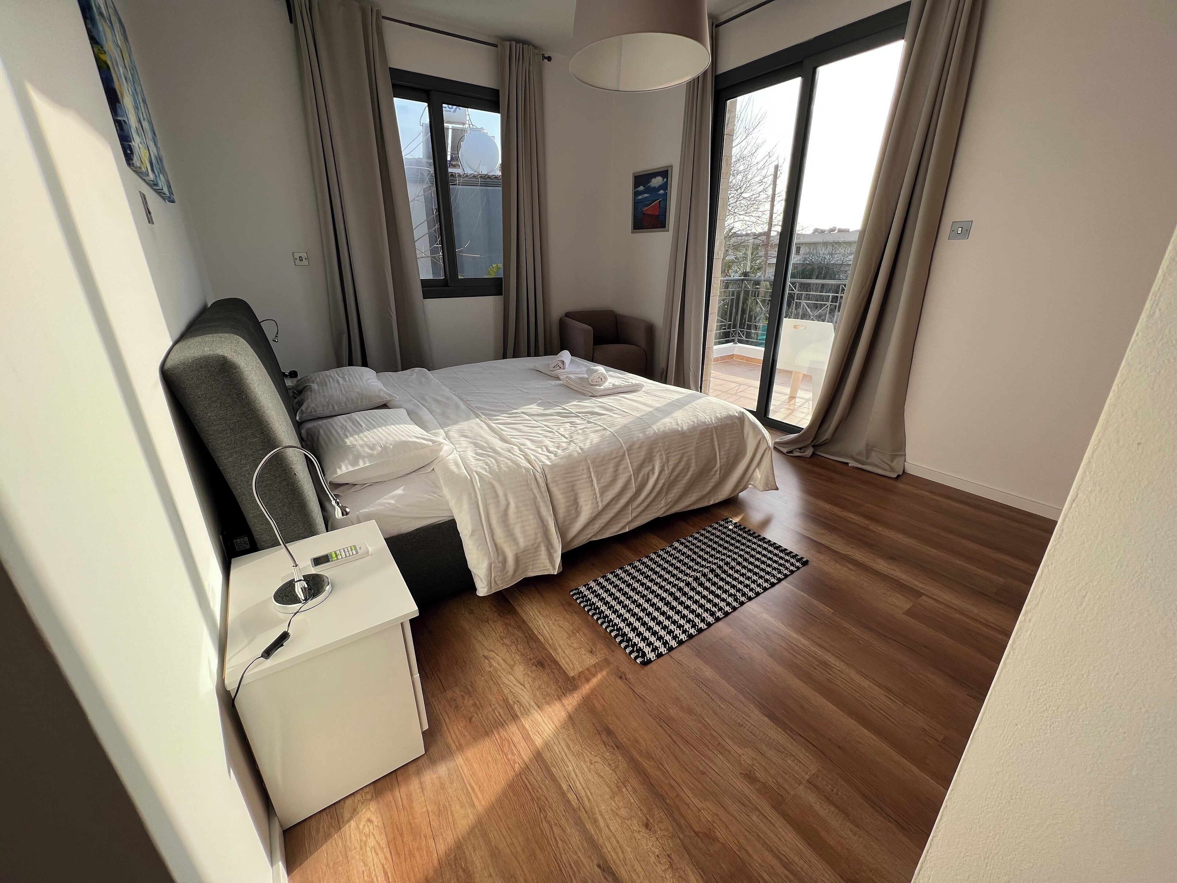A modern bedroom with a large bed, a bedside table with a lamp, a small chair, and a view of the outdoors through a sliding glass door.