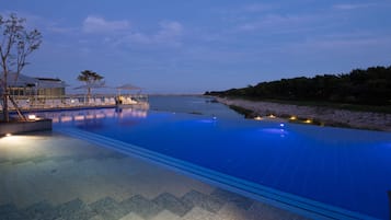 A serene infinity pool with clear blue water, surrounded by lounge chairs and umbrellas, set against a backdrop of trees and a distant shoreline.