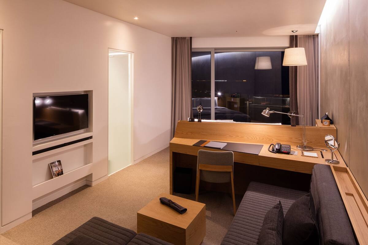 A hotel room with a TV, a desk with a chair, a sofa, and a window with a view of the city at night.
