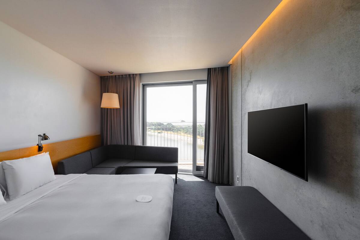 A modern hotel room with a large bed, a sofa, a flat-screen TV, and a balcony with curtains.