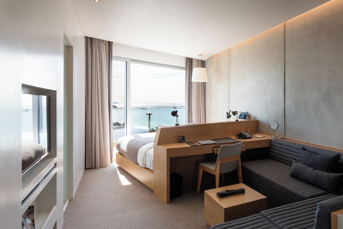 A modern hotel room with a large bed, a desk, a sofa, and a view of the sea.