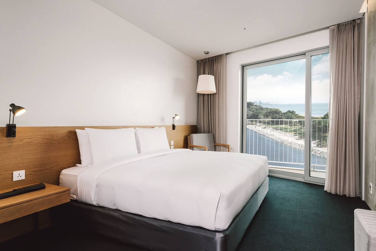 A hotel room with a large bed, a desk, a chair, a TV mounted on the wall, and a balcony with a view of the sea.
