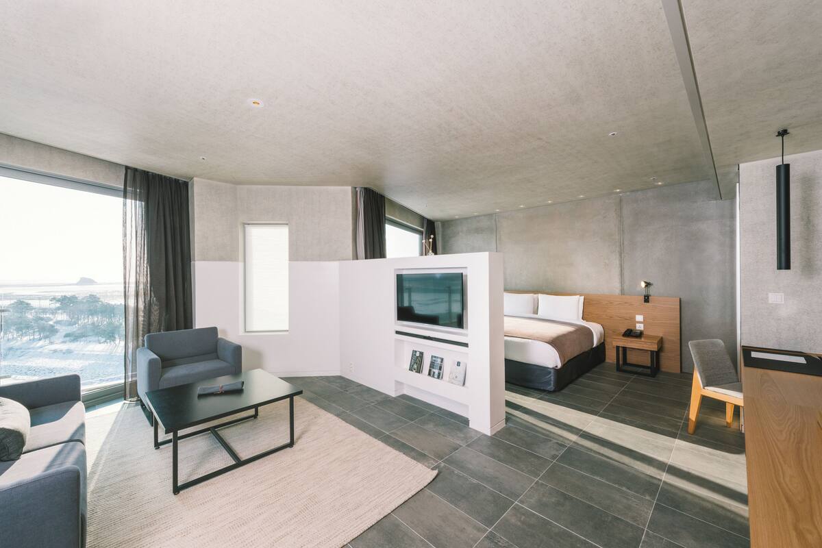 A modern hotel room with a large window, a sofa, a bed, a TV, and a wooden side table.