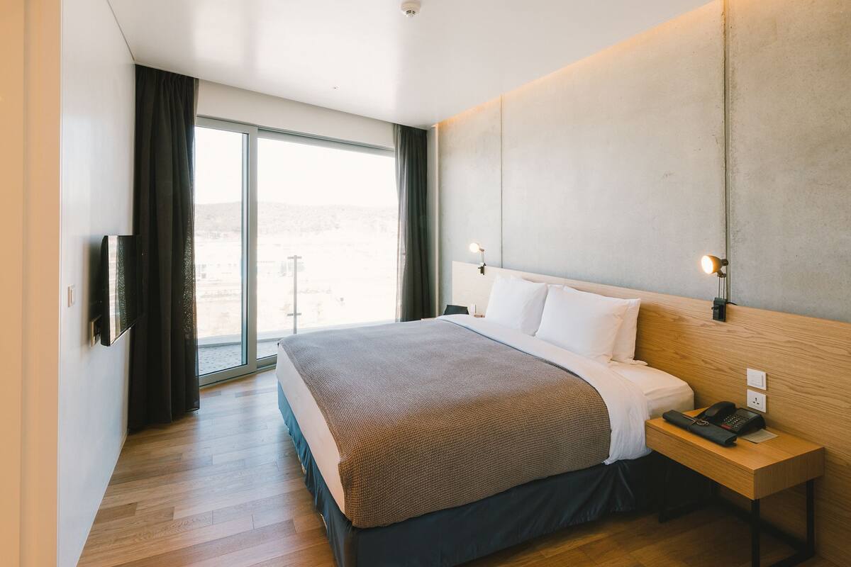 A modern hotel room with a large bed, a wooden nightstand, a telephone, and a view of the outdoors through a large window.