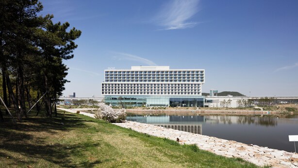 A modern hotel with a glass facade, located near a body of water and surrounded by trees.