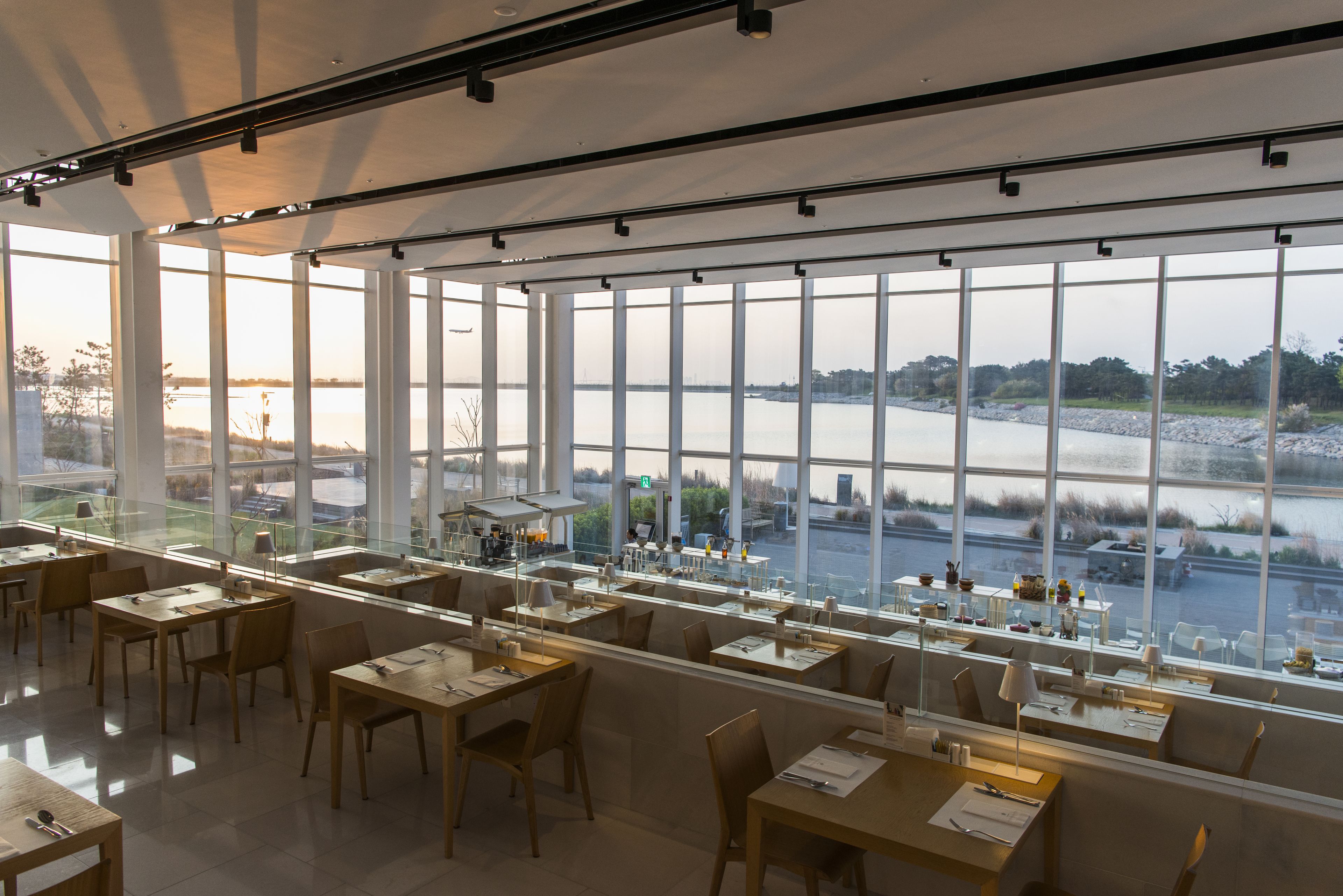 Un restaurante espacioso con amplios ventanales que ofrecen vistas a un río y al paisaje urbano. Las mesas están ordenadas con sillas, y el interior tiene un diseño moderno y limpio.
