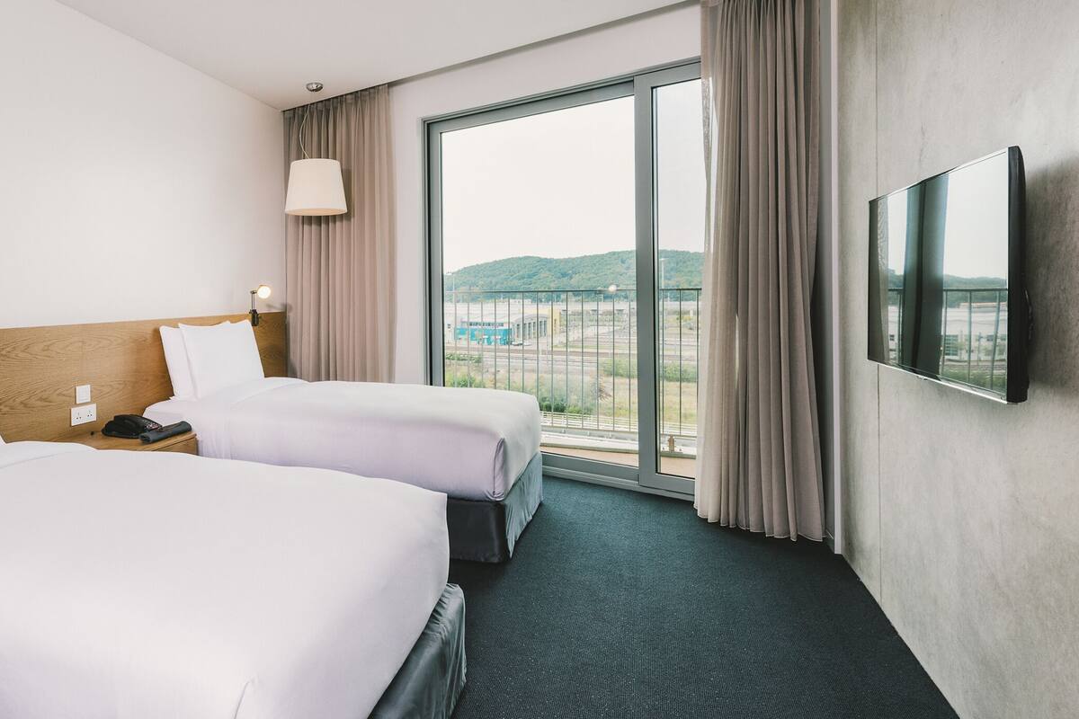A hotel room with two beds, a TV, a lamp, and a large window with a view of a bridge.