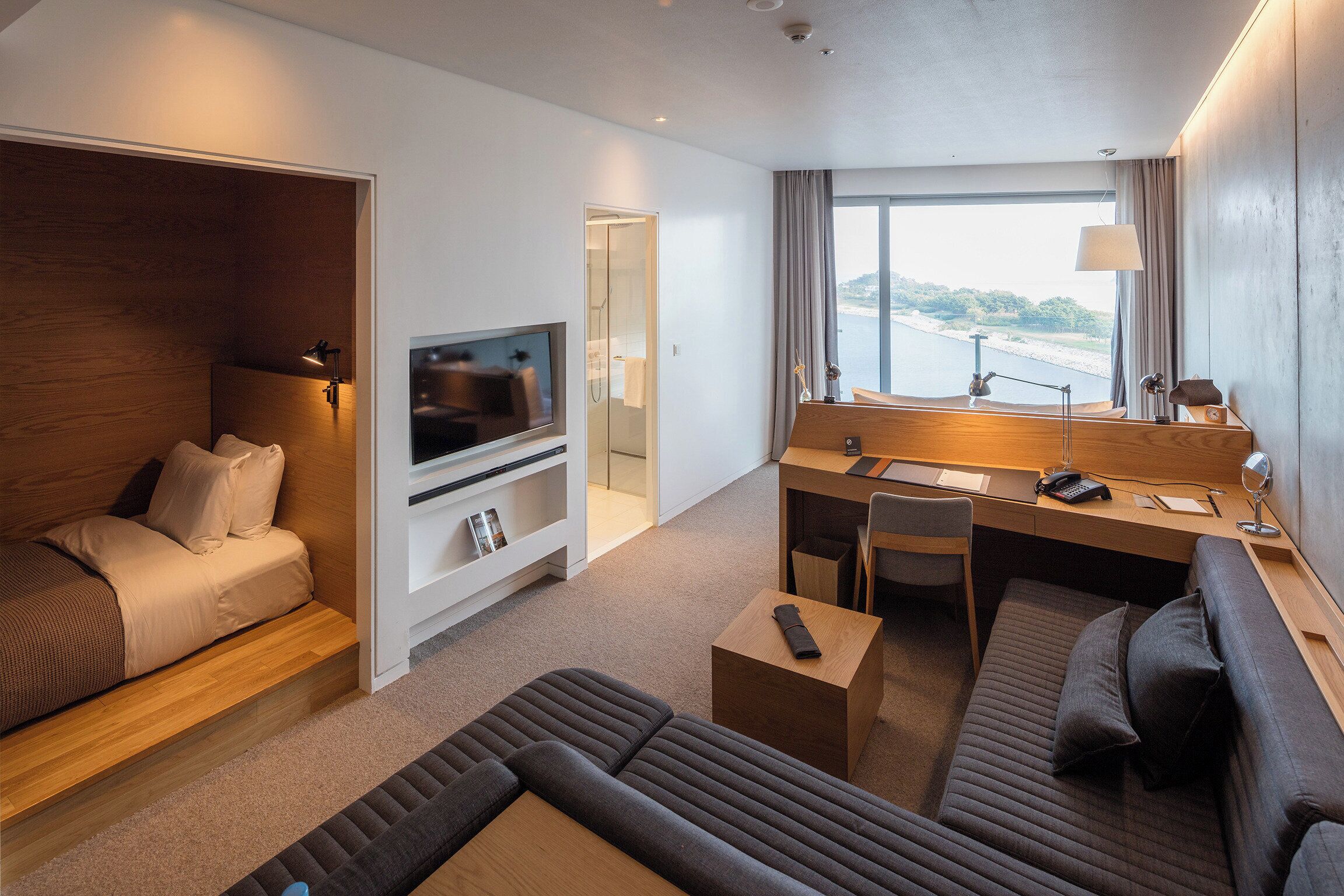 A modern hotel room with a sofa, a small desk, a television, and a large window offering a view of a body of water.