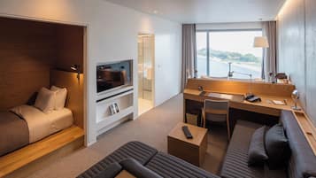 A modern hotel room with a sofa, a small desk, a television, and a large window offering a view of a body of water.