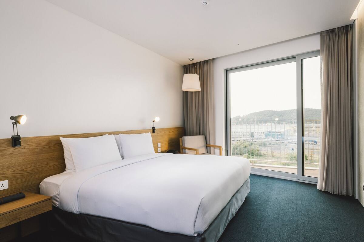 A modern hotel room with a large bed, a flat-screen TV, a desk, and a large window offering a city view.