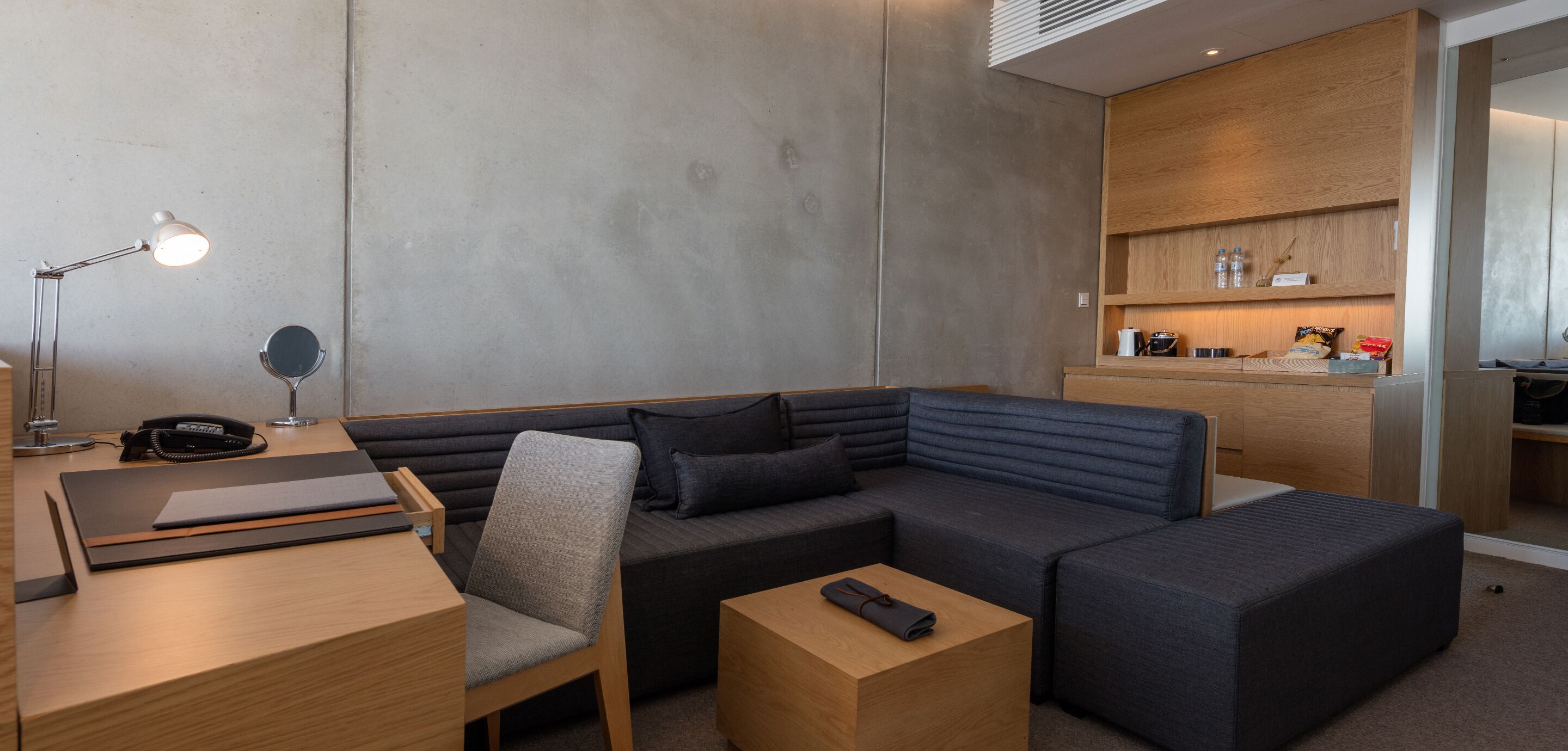 Quarto de hotel moderno com sofá modular, escrivaninha com luminária, telefone e pequena mesa.