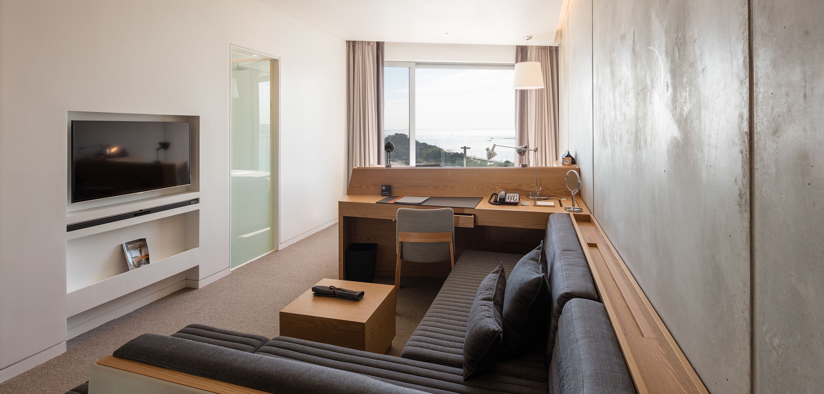 Une chambre d’hôtel moderne dotée d’une télévision à écran plat, d’un bureau avec une chaise, d’un canapé et d’une grande fenêtre donnant sur un paysage agréable.