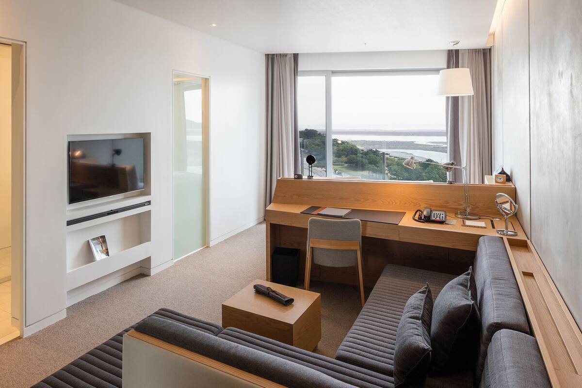 A modern hotel room with a flat-screen TV, a desk with a chair, a sofa, and a large window offering a city view.