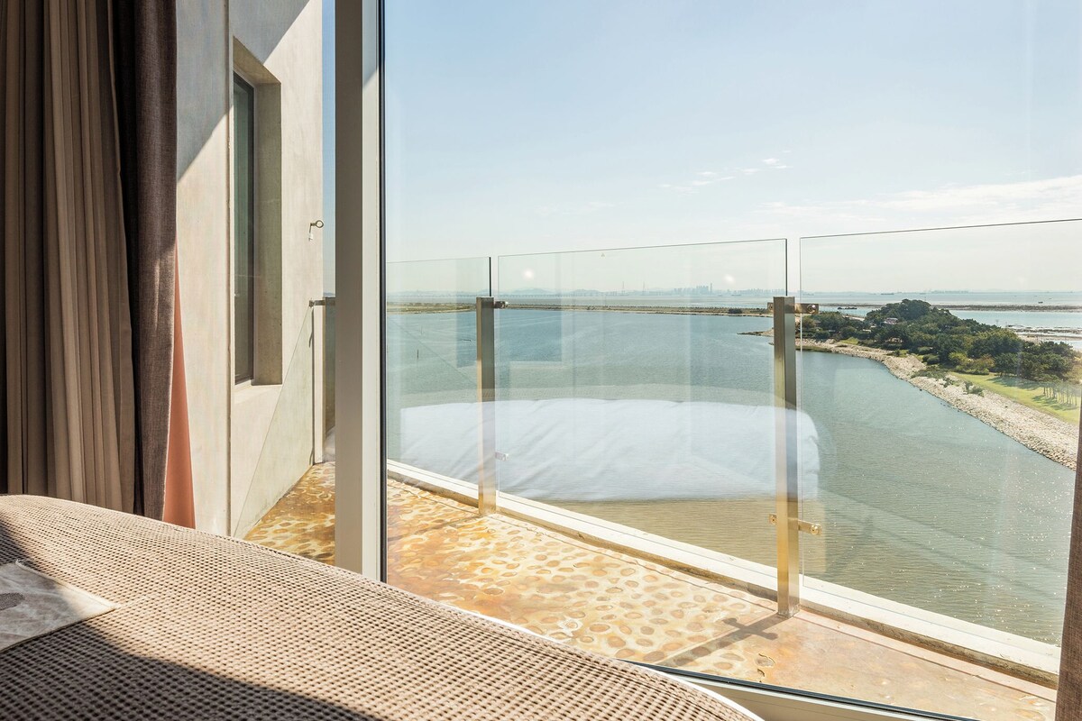 A balcony with a glass barrier, a bed with a white mattress, and a view of a river and distant city skyline.