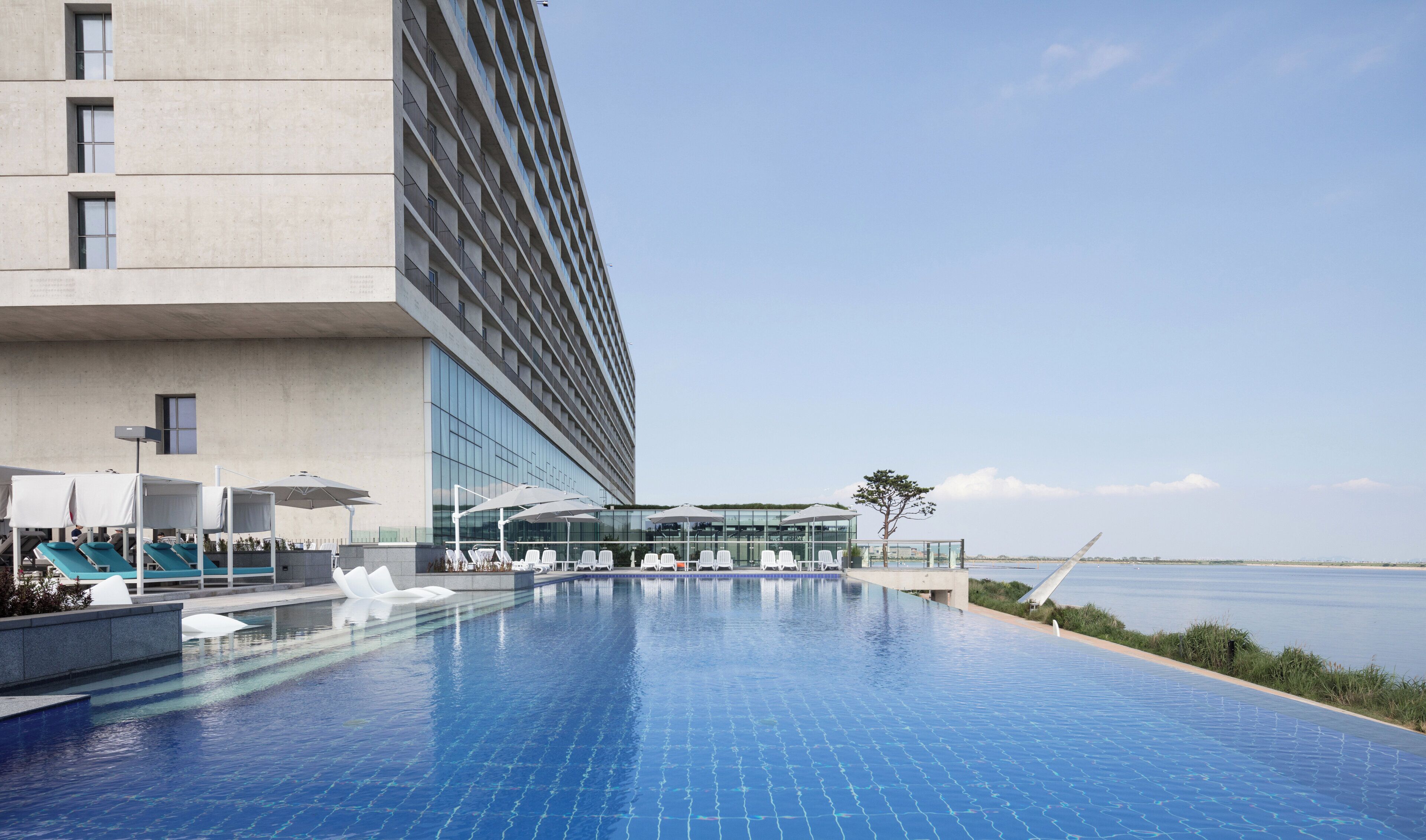 Et moderne hotel med en roftop infinity-pool, liggestole og et glasomkranset område med udsigt over vandet.